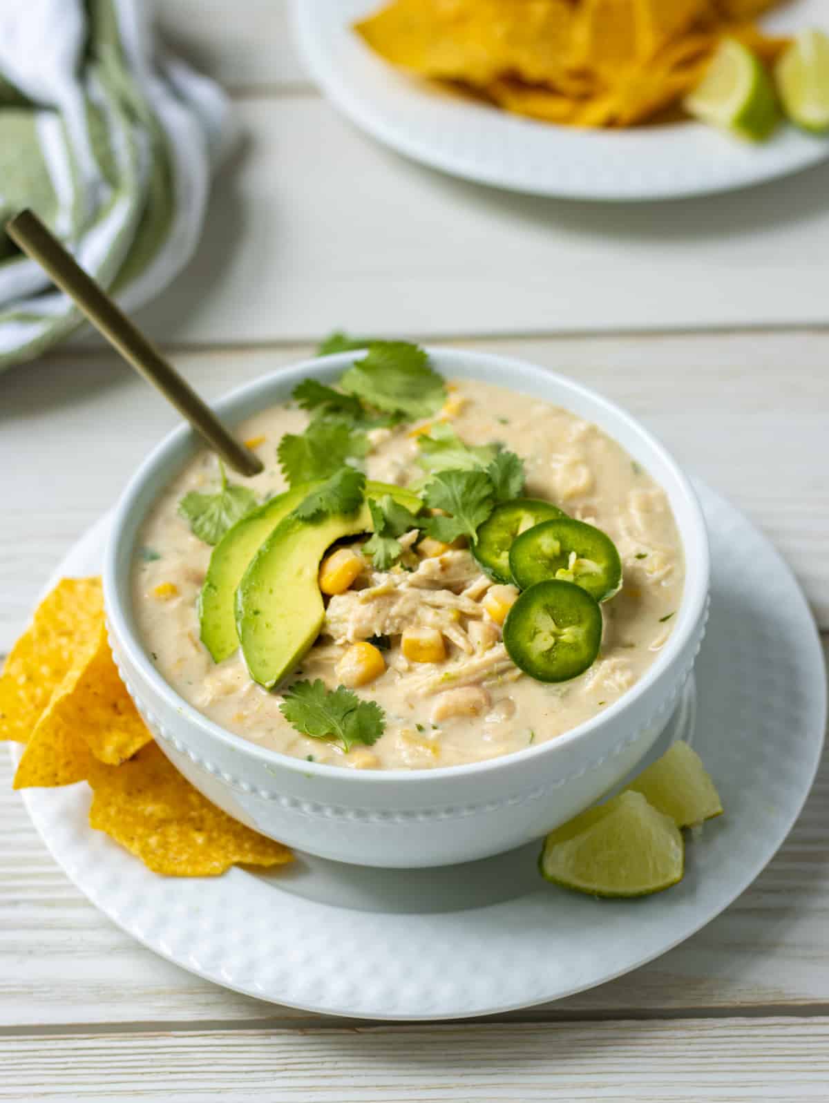 Bowl of white chicken chili topped with sliced avocado, jalapeno, and cilantro on a plate with tortilla chips and lime wedges.