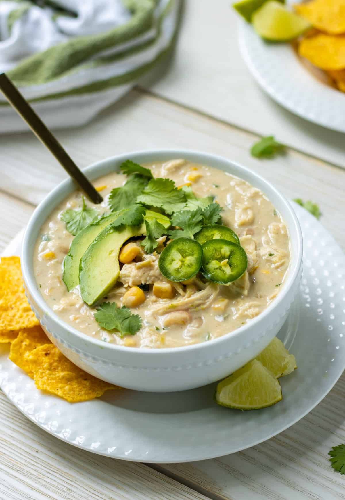 Bowl of white chicken chili topped with avocado, jalapeño, and cilantro on a plate with tortilla chips and lime wedges.
