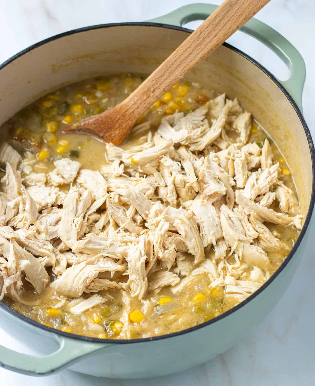 Shredded chicken added back to the pot of white chicken chili with a wood spoon.