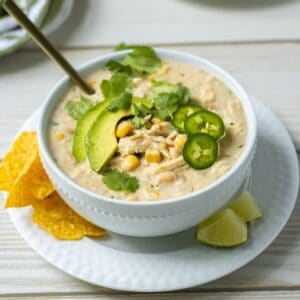 Bowl of white chicken chili on a plate with tortilla chips and lime wedges.