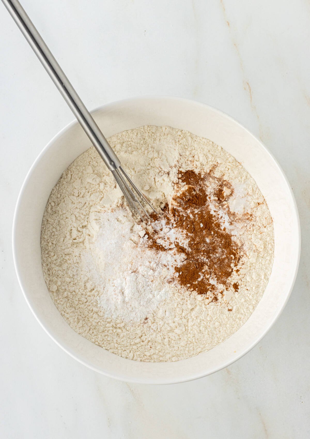 Flour, baking soda, baking powder, salt, and cinnamon in a bowl with a whisk.