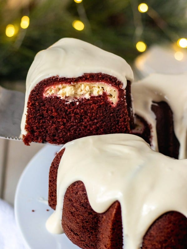 Red velvet bundt cake with frosting on a cake stand with Christmas lights and pine branches in the background.