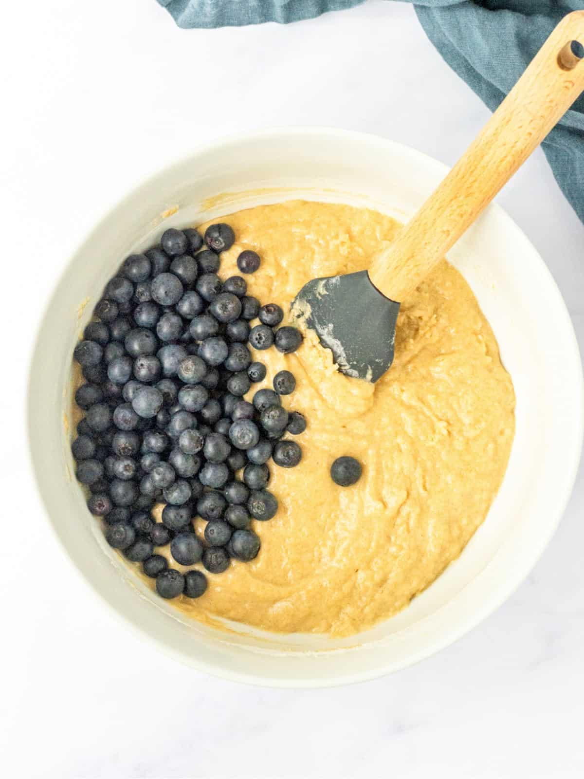 Blueberries on top of muffin batter in a bowl with a spatula.