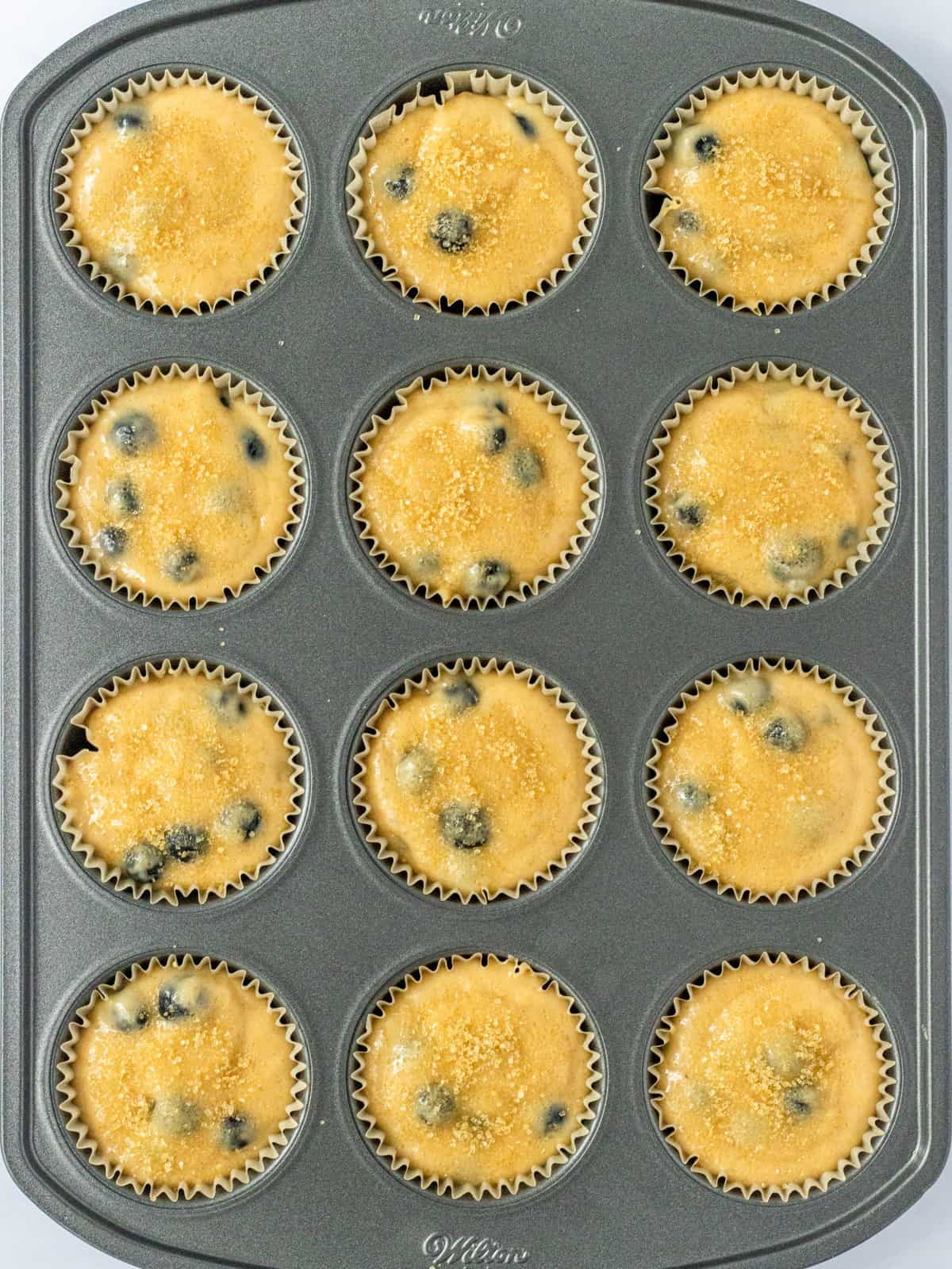 Blueberry muffin batter-filled liners in a muffin pan topped with coarse sugar.