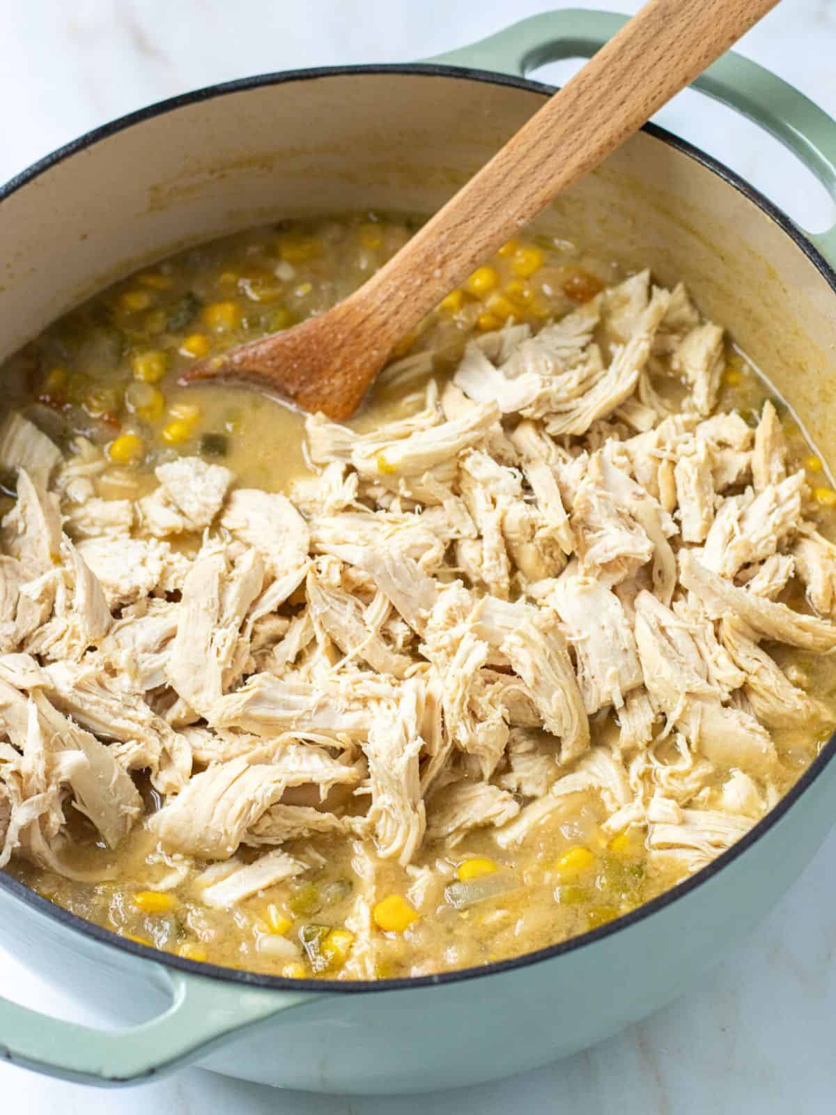 Shredded chicken added back to broth in Dutch oven with a wood spoon.